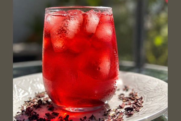 SoGo Pure Hibiscus Lavender iced tea on a sunny day, served on a white tray with loose hibiscus lavender tea surrounding a glass filled with ice cubes and rich red tea, with beads of condensation on the exterior.