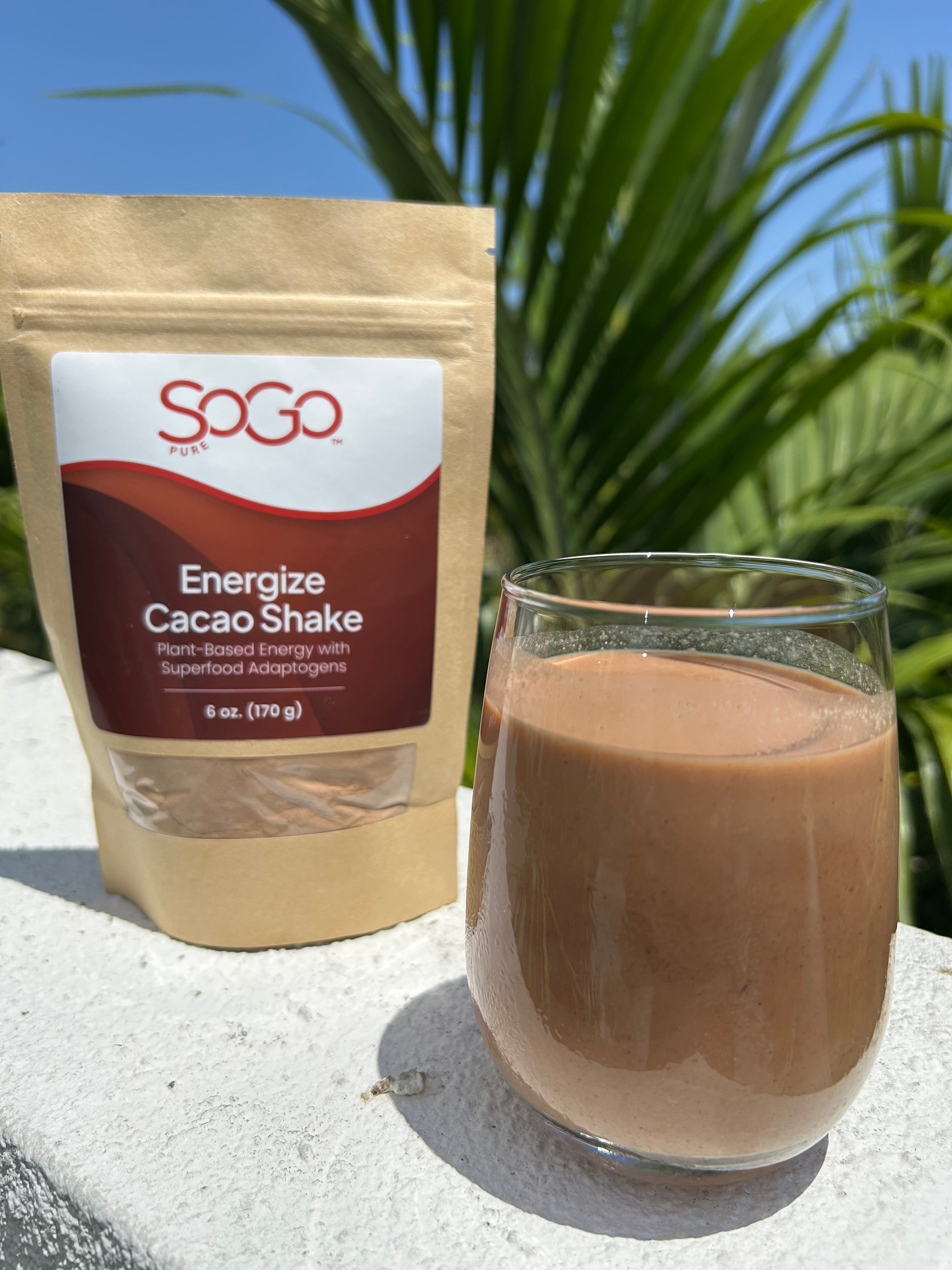 SOGO Energize Cacao Shake Powder package and glass of cacao shake on a white ledge with palm leaves in the background on a sunny day.