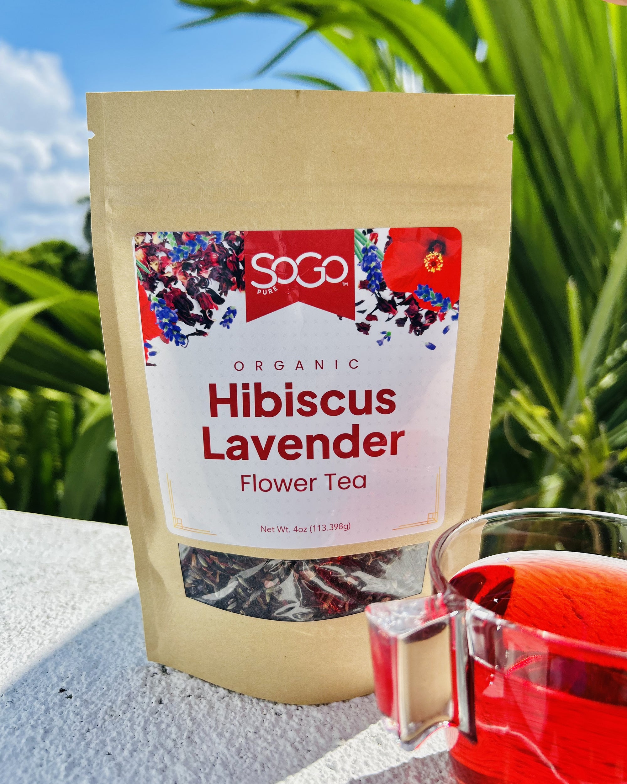 SoGo Pure Organic Hibiscus Lavender Flower Tea package beside a cup of hibiscus lavender tea, set against tropical palms, blue sky, and white clouds in a sunny outdoor setting. The tea has a soft red color.