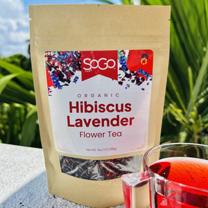 SoGo Pure Organic Hibiscus Lavender Flower Tea package beside a cup of hibiscus lavender tea, set against tropical palms, blue sky, and white clouds in a sunny outdoor setting. The tea has a soft red color.