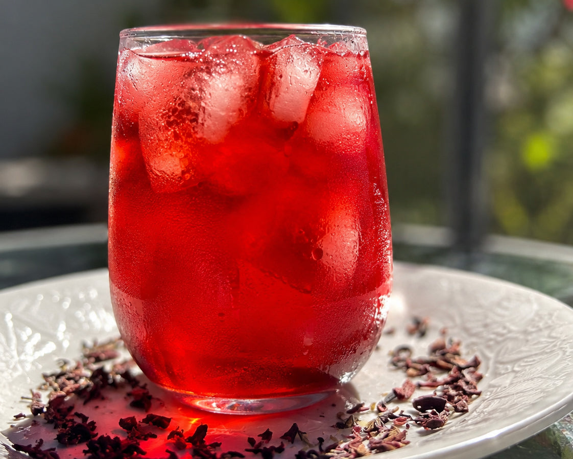 Glass of red hibiscus lavender iced tea with hibiscus and lavender flowers on a white plate outdoors.