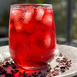 Glass of red hibiscus lavender iced tea with hibiscus and lavender flowers on a white plate outdoors.