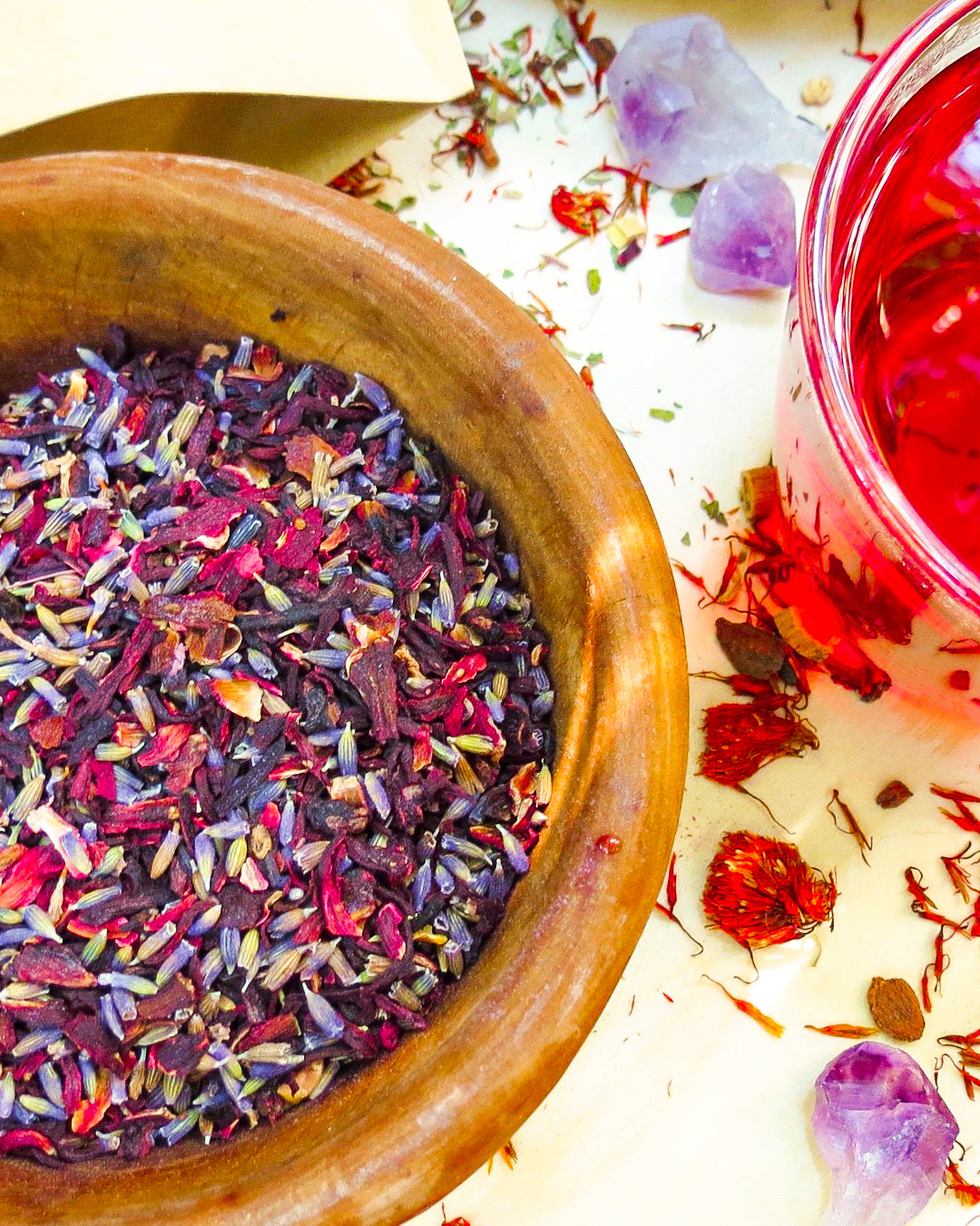 SoGo Pure Organic Hibiscus Lavender Flower Tea in a wooden bowl filled with loose hibiscus petals and lavender flowers, with a cup of red herbal tea. On the table are dried flowers and amethyst crystals.