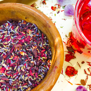 SoGo Pure Organic Hibiscus Lavender Flower Tea in a wooden bowl filled with loose hibiscus petals and lavender flowers, with a cup of red herbal tea. On the table are dried flowers and amethyst crystals.